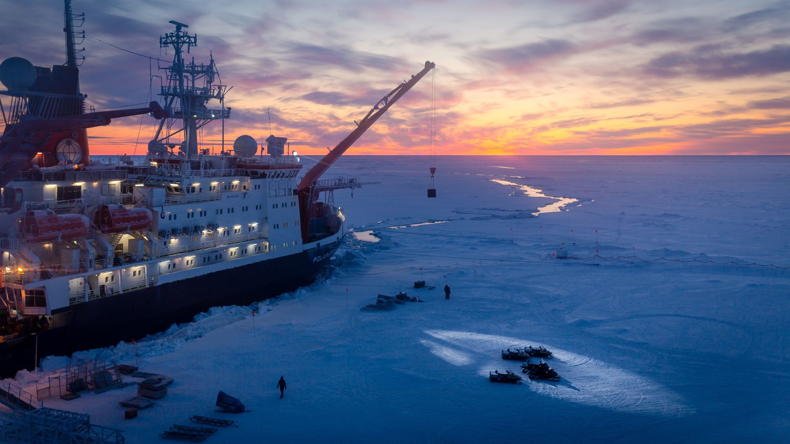 A large icebreaker ship is frozen in the sea ice, with various people and equipment on top of the surrounding ice.